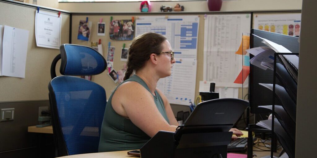 Woman in a cubicle working on a computer, surrounded by office materials, family photos, and charts on the wall, at Chadwick Optical.