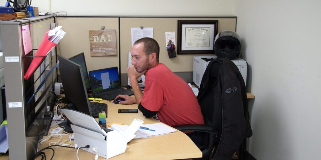 Man in a red shirt focused on his computer at a cubicle desk, with a motorcycle helmet and framed diploma on the wall behind him.