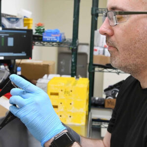 lab-staff-10 Bryan examines a lens in the Quality Control area of Chadwick Optical, wearing blue gloves and focused on the inspection, with lab equipment and storage boxes in the background.