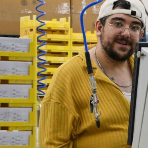lab-staff-11 Matt, standing near stacked trays and a monitor in Chadwick Optical’s lab, smiles toward the camera, with a blue air hose coiled overhead, part of the equipment setup in the workspace.