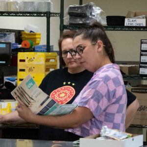 lab-staff-12 Jenn and Jordan examine a catalog together in Chadwick Optical’s lab area, surrounded by organized supplies and equipment, likely discussing product options or inventory.
