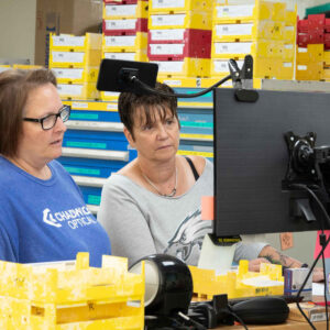 lab-staff-13 Jenn (left) and Susan (right) examine a computer screen together in Chadwick Optical’s lab, surrounded by trays of organized lenses, discussing work-related details.