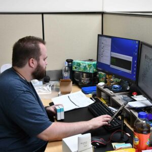 leadership-staff1 Alex at Chadwick Optical seated at his desk, working on a computer with a dual monitor setup, surrounded by office items and various personal desk decorations.