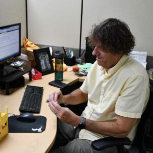 leadership-staff2 Tom at Chadwick Optical seated at his desk, working with small parts in his hands, with his computer and office items arranged on the desk around him.