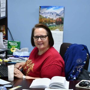 leadership-staff9 Jenn sitting at her desk with open books and documents, a calendar on the wall, working at Chadwick Optical.