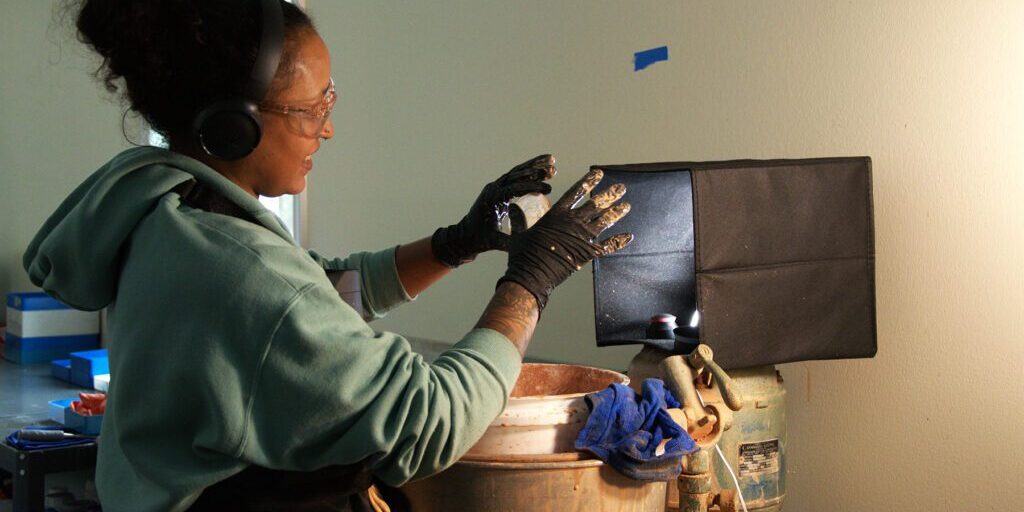 Woman wearing safety goggles and gloves working on a lens casting process under focused lighting, with hands positioned over casting equipment at Chadwick Optical.