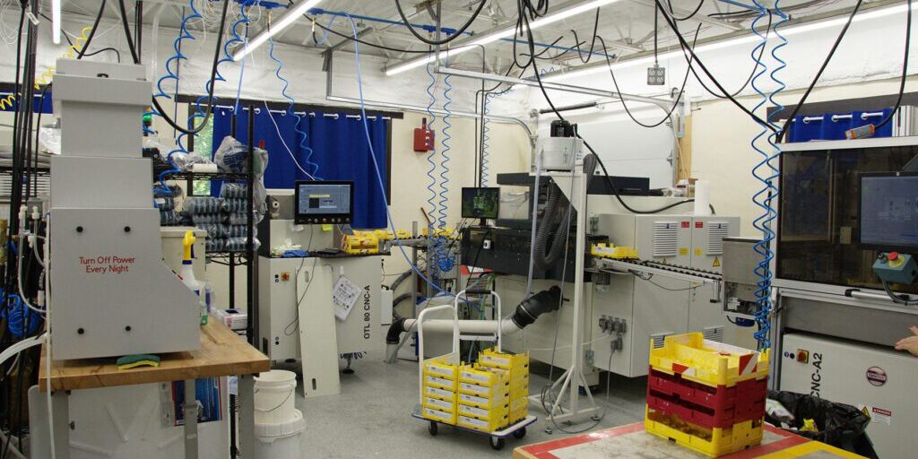 Industrial lab setup with multiple machines, hanging cables, and yellow storage bins, showcasing a manufacturing environment.