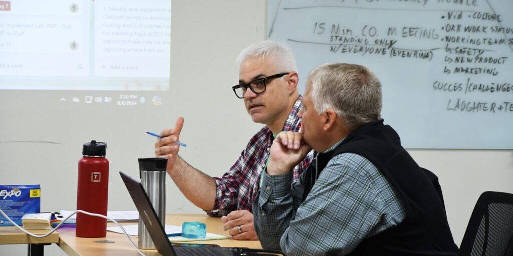 Two men engaged in a discussion during a meeting at a conference table, with notes visible on a whiteboard in the background.