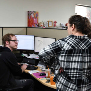 office-staff-10 John and Amanda at Chadwick Optical, engaged in discussion at John's desk workstation, with dual monitors displaying work documents.