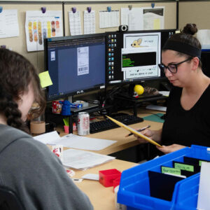 office-staff-5 Faith and Jordan working together at a desk, with a computer screen showing shipping labels and workspace materials surrounding them.