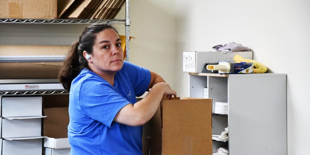 Woman in a blue shirt, wearing an earbud, packing a cardboard box in a workspace with shelves and packing materials.