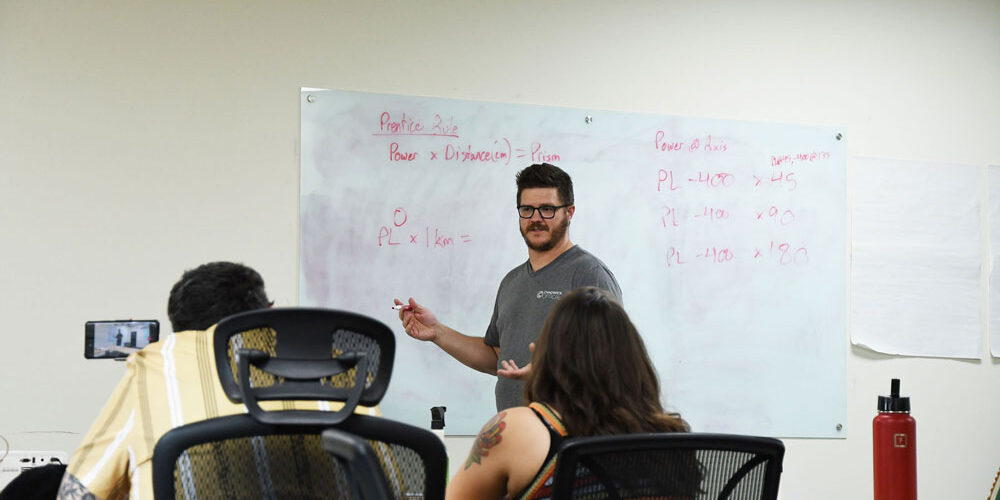Charlie leads a training session at Chadwick Optical, explaining equations on a whiteboard to a group of engaged employees.
